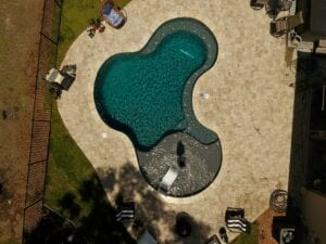 High angle drone view of a kidney-shaped backyard swimming pool with dark plaster, a wide tanning ledge, and tan paver decking surrounded by a black fence