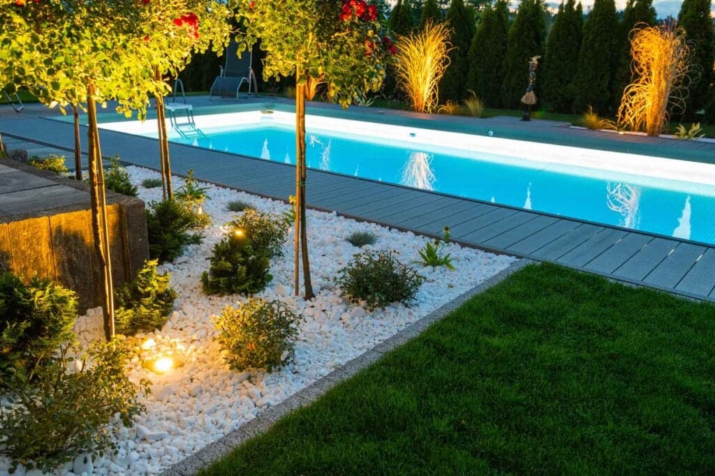 Modern, narrow lap swimming pool at night with bright blue underwater LED lighting, surrounded by a gray composite deck, illuminated landscaping with white river rock, and manicured trees.