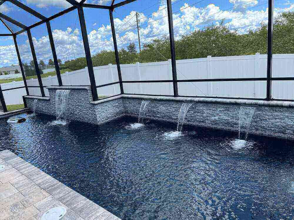 Custom pool wall with stacked stone veneer and four sheer descent waterfalls, set inside a black screen enclosure with a dark pool interior and surrounding paver deck.