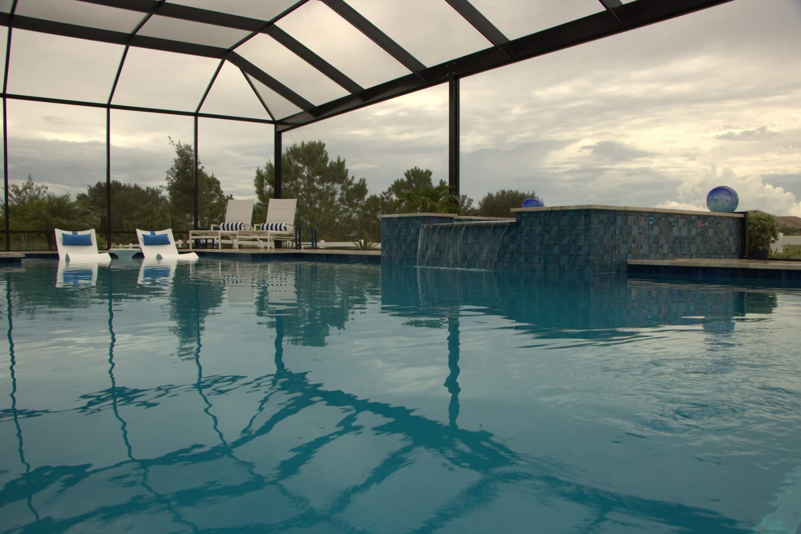 Water-level view of a swimming pool inside a black screen enclosure, featuring a raised blue mosaic tile water wall with small waterfalls and submerged white lounge chairs against a cloudy sky.
