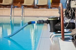 A portable black pool cleaning pump system with a blue suction hose submerged in a clear outdoor swimming pool at a resort.