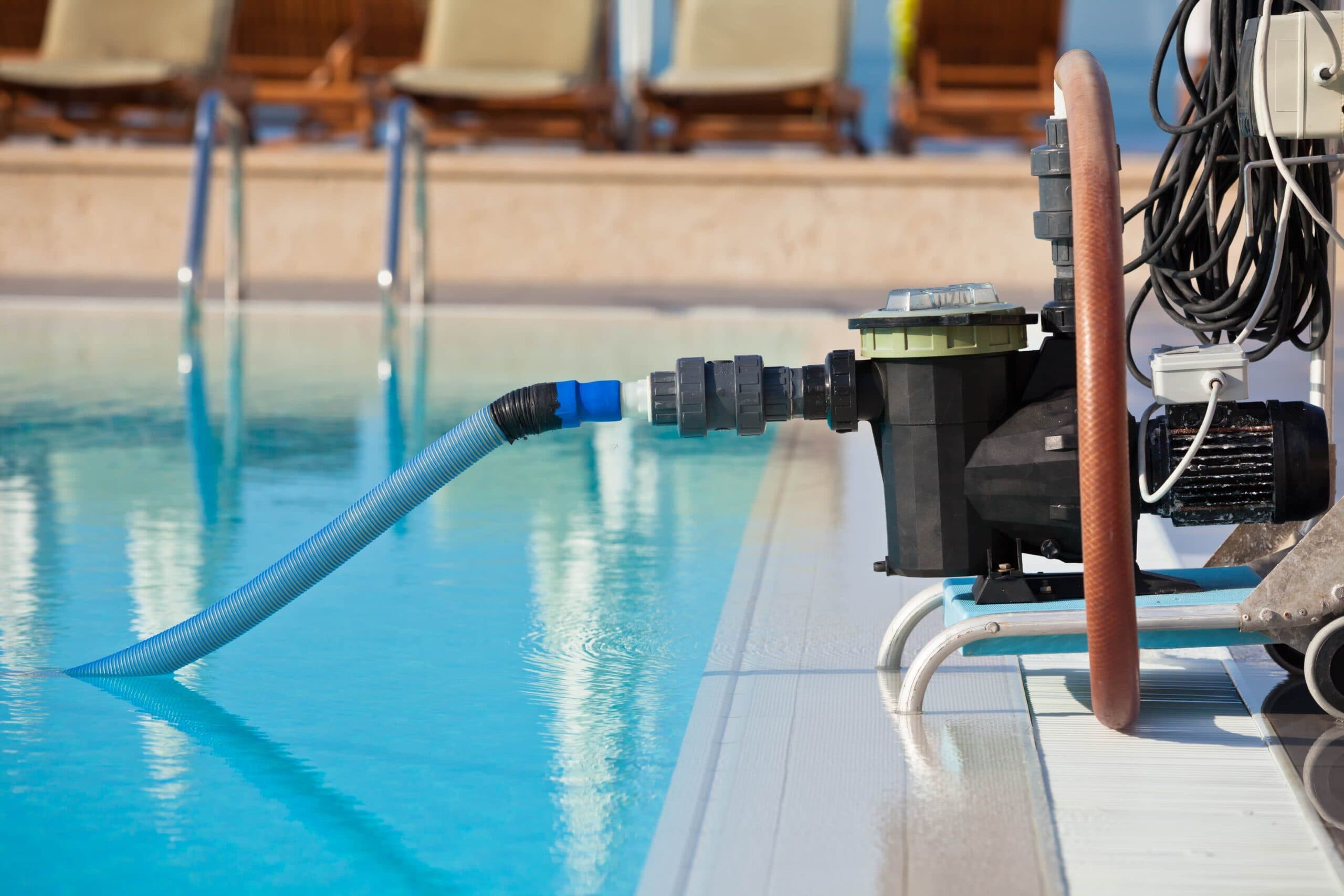 A portable black pool cleaning pump system with a blue suction hose submerged in a clear outdoor swimming pool at a resort.