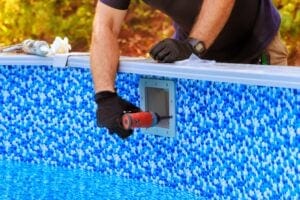 A close-up of a pool technician's hands in black gloves using a power drill to secure a skimmer faceplate onto a blue mosaic-patterned vinyl pool liner.
