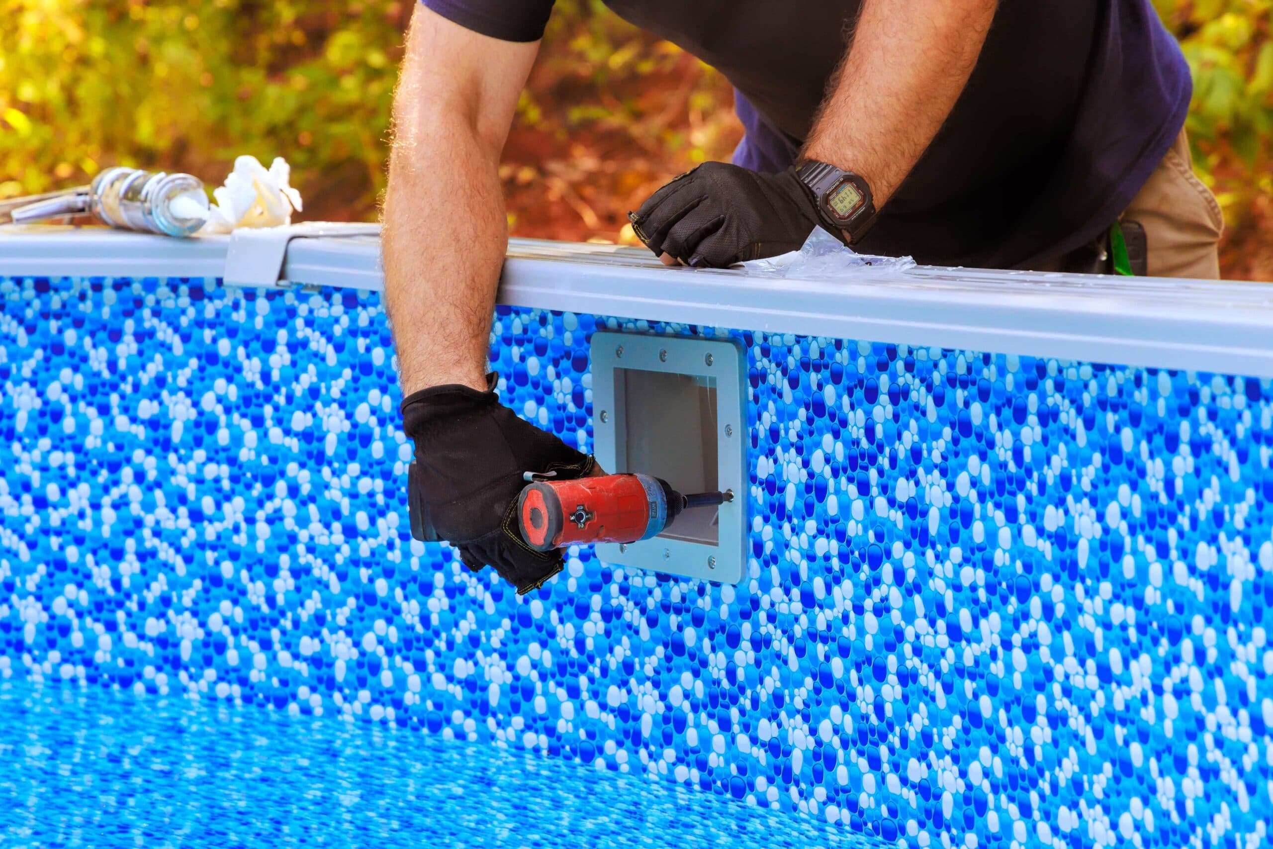 A close-up of a pool technician's hands in black gloves using a power drill to secure a skimmer faceplate onto a blue mosaic-patterned vinyl pool liner.