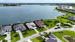 High-angle drone photograph of a row of modern luxury white-walled mansions with grey roofs bordering a large calm lake with private docks and manicured green lawns.