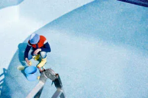 A worker wearing yellow boots and a hat crouches in an empty, blue-tiled swimming pool, using a sponge and a blue bucket to clean the tiles manually.