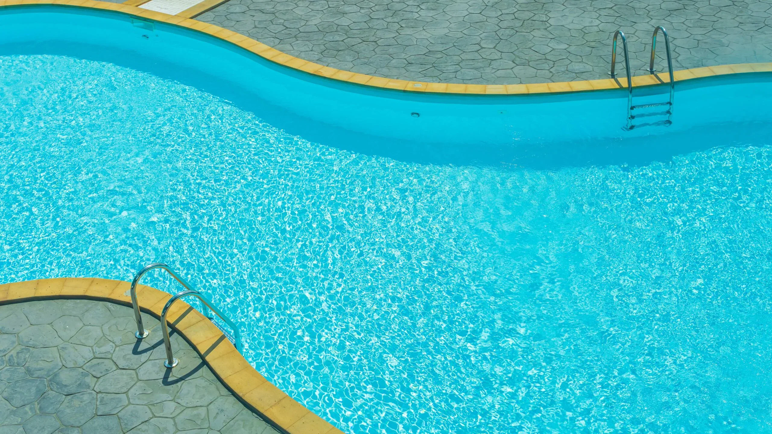 An inviting, winding turquoise swimming pool with sparkling clear water, metal ladders, and a grey patterned stone deck at a resort.