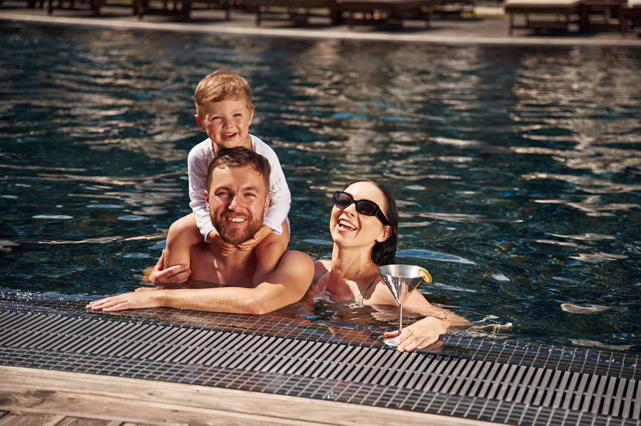 A smiling father with a young son on his shoulders and a mother holding a cocktail, enjoying a sunny day in a luxury outdoor swimming pool.