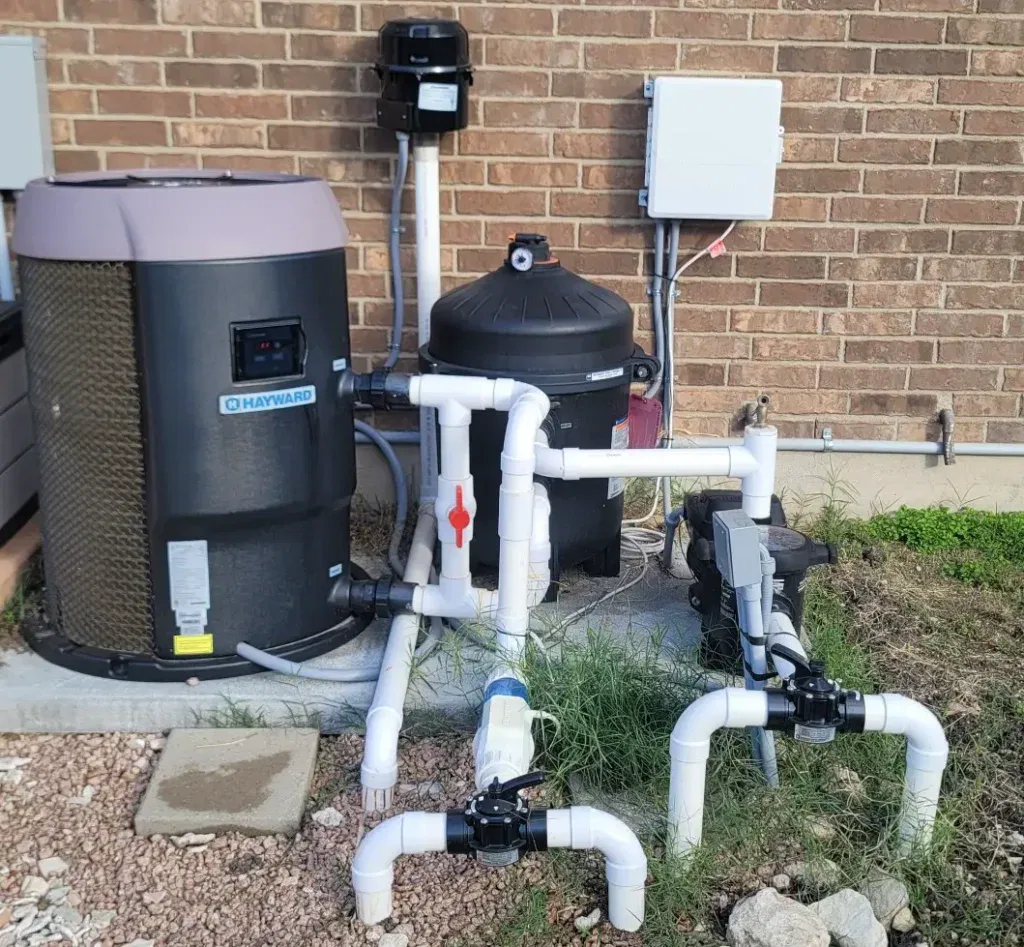 A professional residential pool equipment pad installation featuring a Hayward HeatPro heat pump, a black cartridge filter, and PVC plumbing with Jandy valves against a brick exterior wall.
