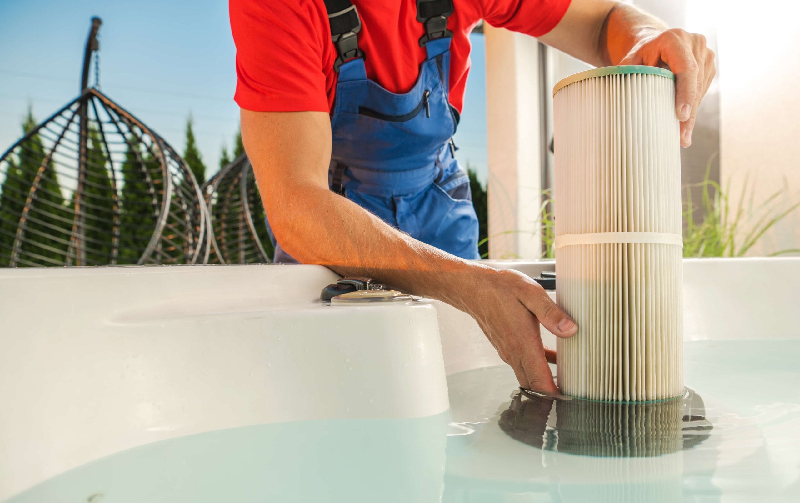Close-up of a service technician in blue overalls inserting a new white pleated cartridge filter into the filter housing of a hot tub filled with clear water.