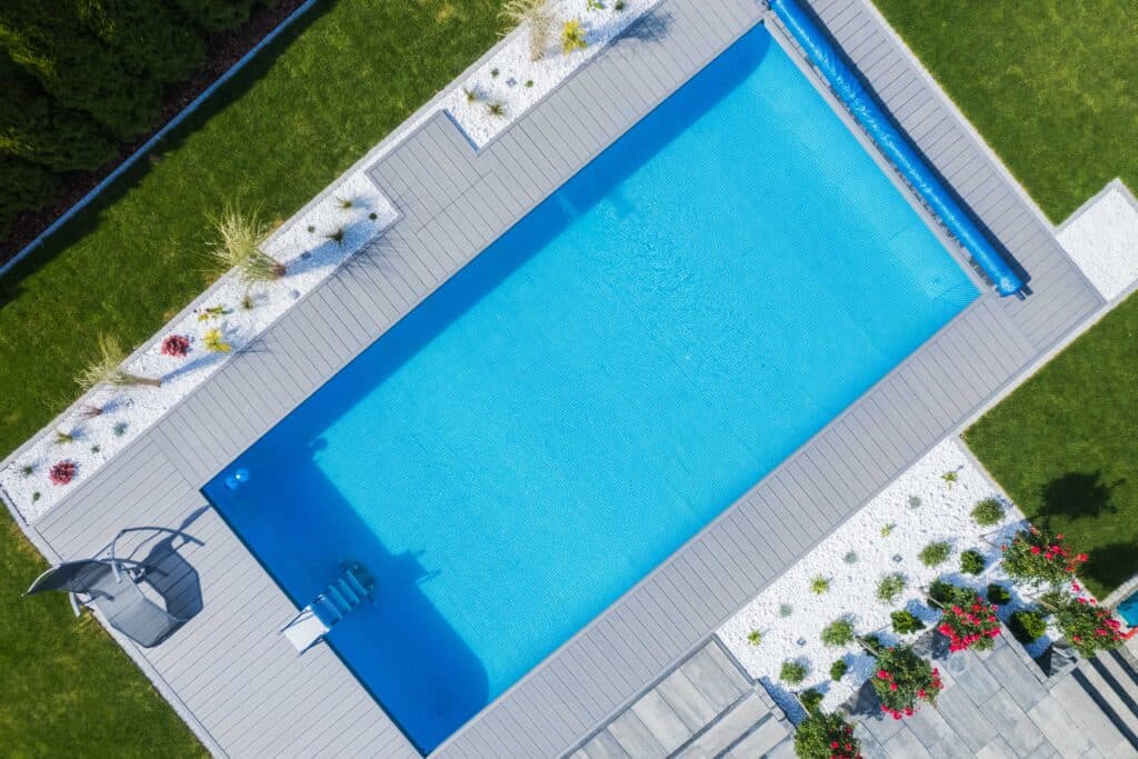 Aerial view of a modern rectangular swimming pool with bright blue water, grey composite decking, white gravel landscaping, and a manicured green lawn.