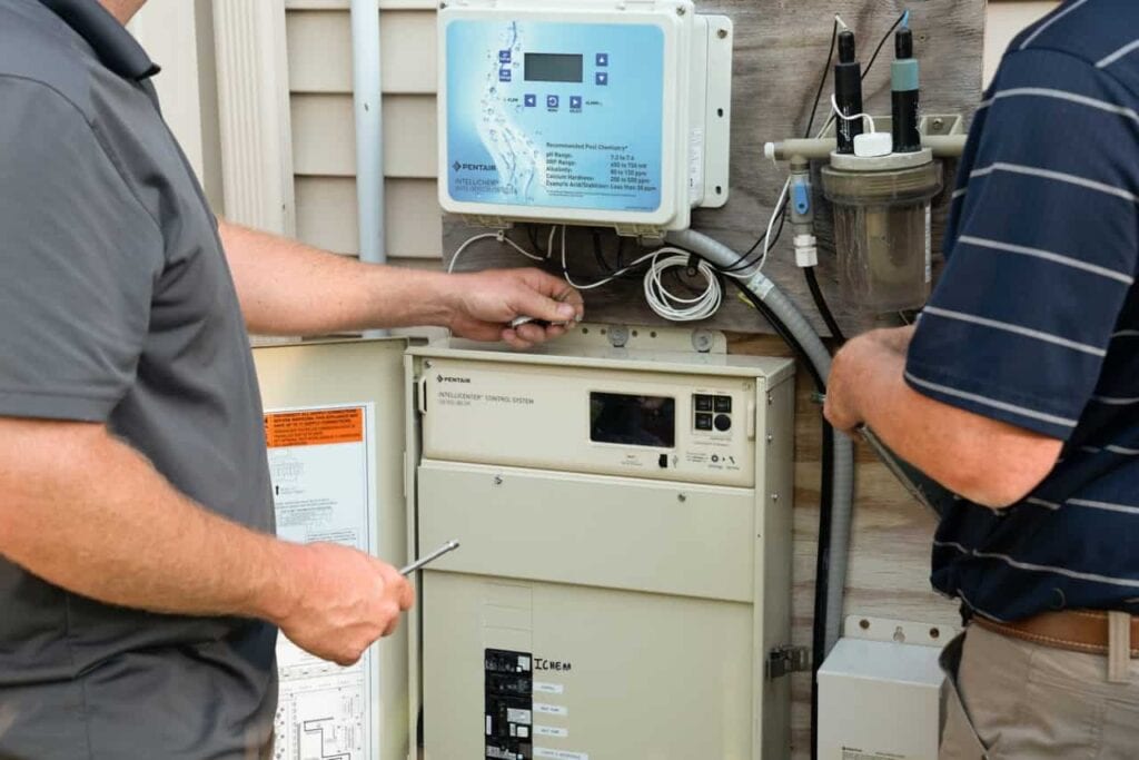Two technicians installing and configuring a Pentair IntelliCenter Control System and IntelliChem Water Chemistry Controller on an outdoor residential pool equipment wall.