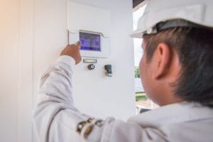 Close-up view of a clean energy technician wearing a white hard hat and work jacket, pointing at and interacting with a touchscreen Human Machine Interface (HMI) panel for a solar power inverter system at a solar farm facility.