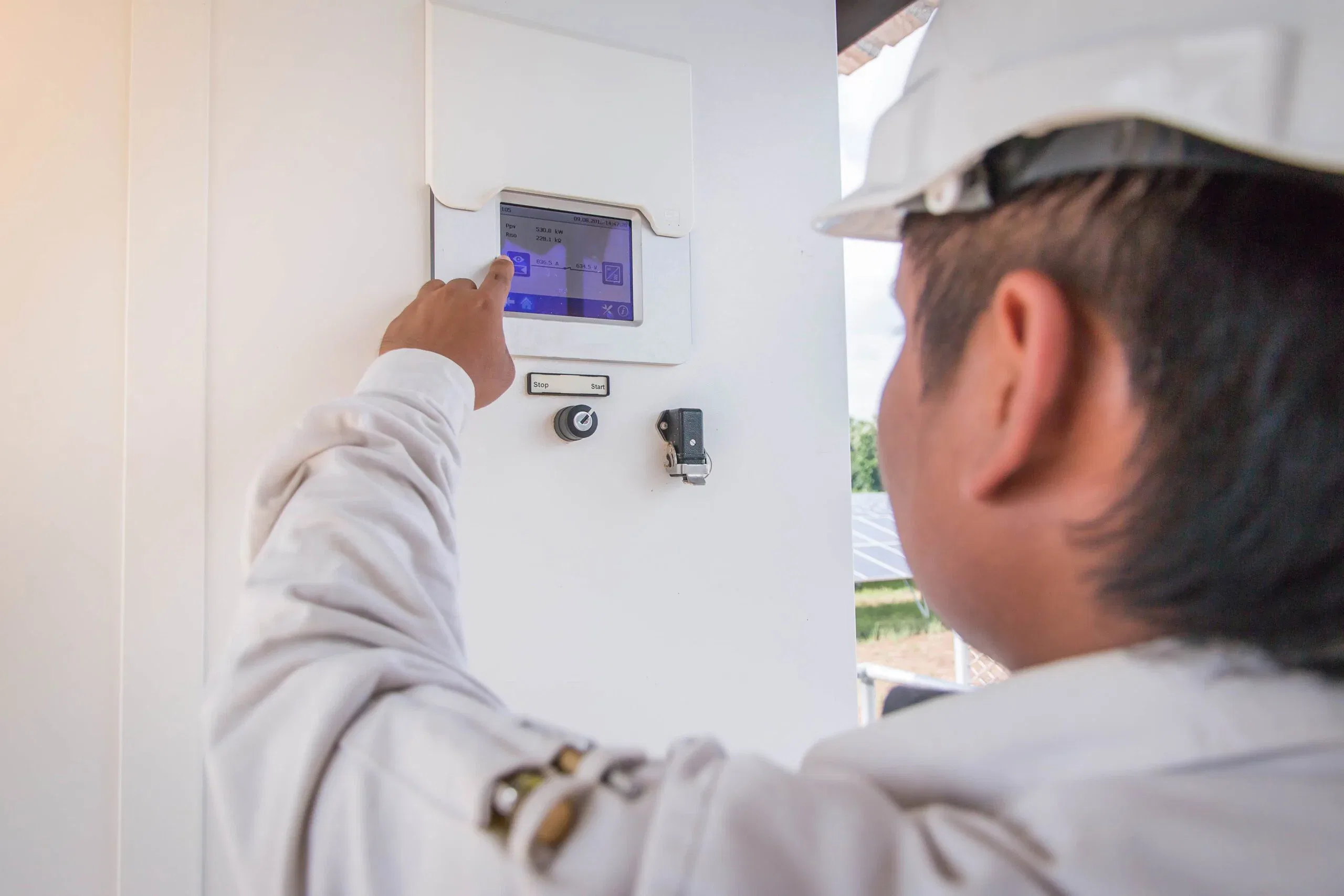Close-up view of a clean energy technician wearing a white hard hat and work jacket, pointing at and interacting with a touchscreen Human Machine Interface (HMI) panel for a solar power inverter system at a solar farm facility.