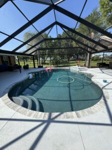 Large freeform swimming pool with dark bottom, rounded tiled steps, and a black-framed screen enclosure on a sunny day