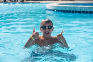 A happy boy wearing swimming goggles gives a thumbs up while playing in a pool during swimming lessons in Phoenix, AZ.